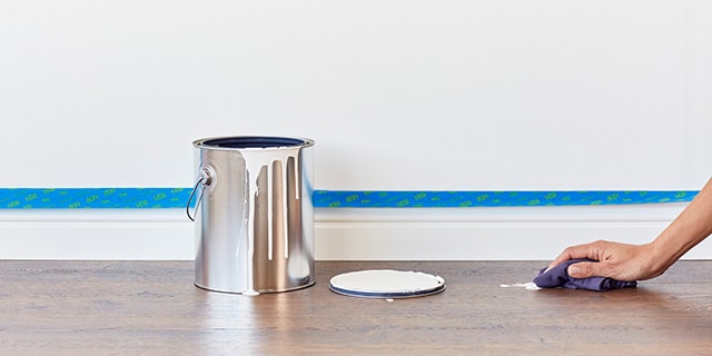 A person wiping paint spilled on a wood floor beside a can of paint.
