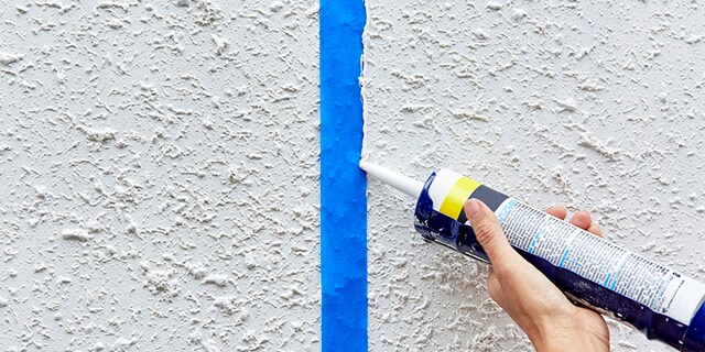 A person applying a thin strip of caulk to the edge of painter's tape on a wall.
