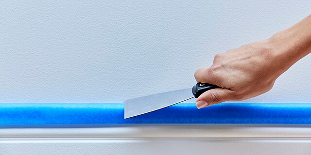 A woman securing tape to a baseboard using a putty knife.

