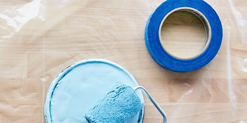 Un rouleau à peinture et un rouleau de ruban pour peintres sur une feuille de plastique étendue sur le plancher.
