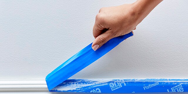 A person removing painter's tape from a baseboard.
