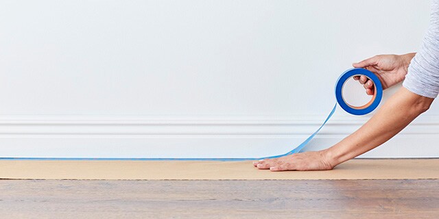 A person using painter's tape to secure rosin paper along a baseboard.
