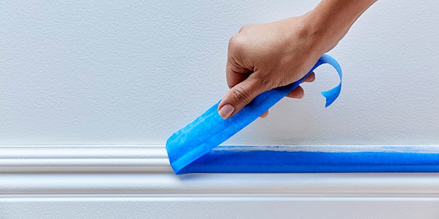 A person removing painter's tape from a baseboard.

