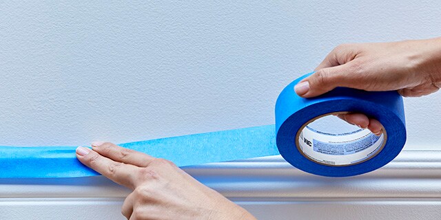 Une main qui applique du ruban pour peintres sur une plinthe.
