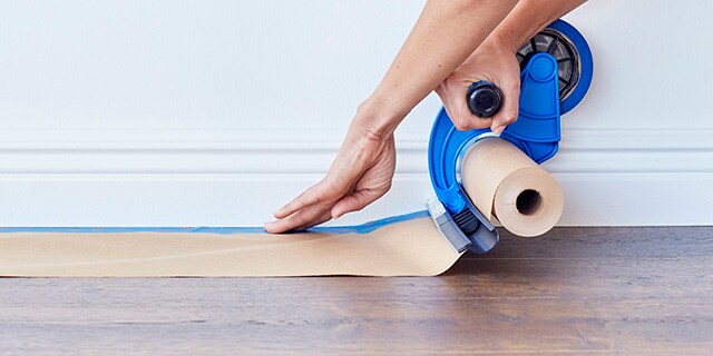 Une femme qui utilise le dévidoir de ruban et de papier pour peintre ScotchBlue(MC) pour protéger un plancher.
